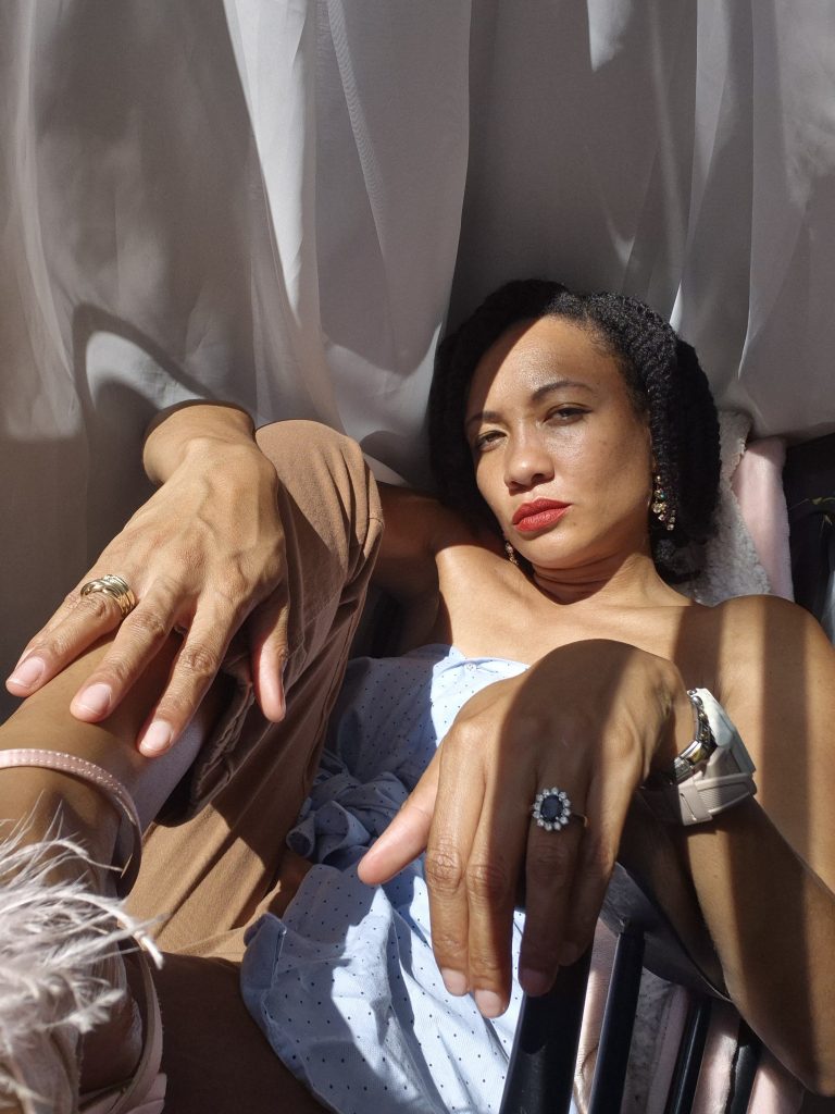 A person lounging in a chair with legs crossed, wearing a light blue top and looking thoughtfully at the camera, surrounded by soft white curtains and natural light.