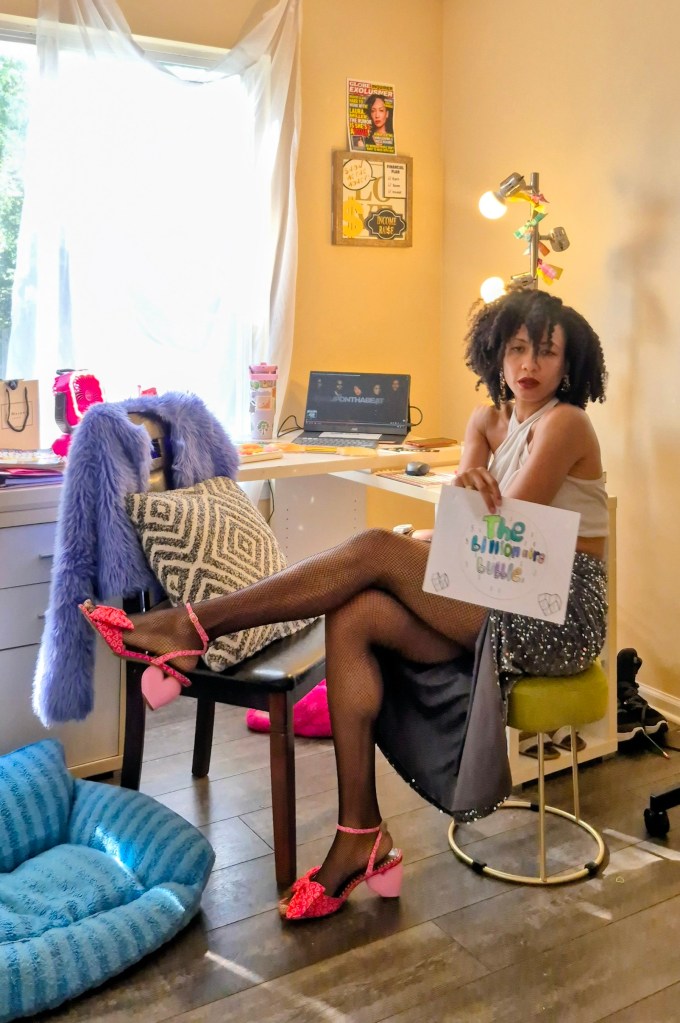 A woman with curly hair sits in a home office, wearing a light-colored top and a flowing skirt. She poses with one leg crossed over the other, showcasing her sparkly pink high heels with heart-shaped accents. In one hand, she holds a sign that says 'The Billionaire Bubble'. The background features a desk with a laptop, various items, and colorful decorations.
