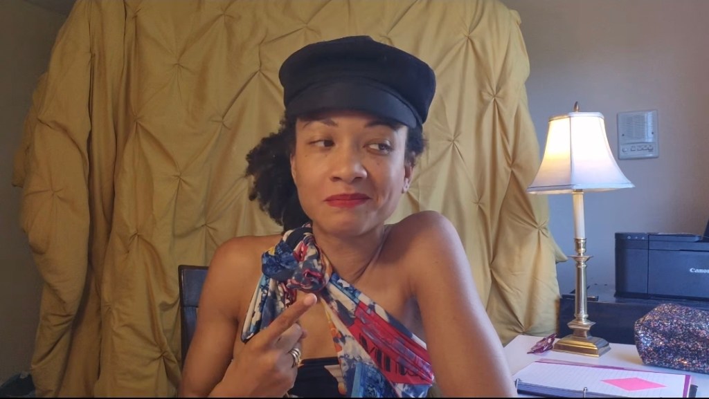 A woman with curly hair wearing a black cap and a colorful top, sitting in front of a lamp and a printer, with a patterned background.